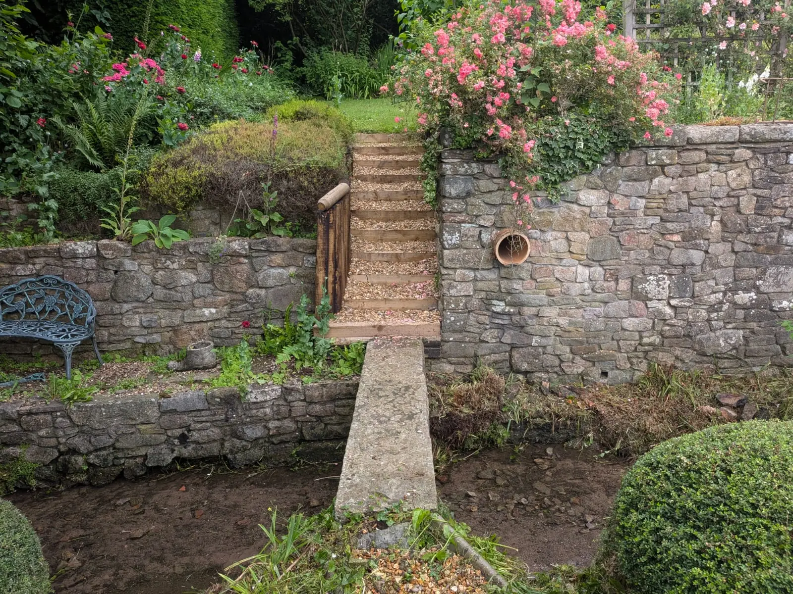 Dry stone retaining walls on two levels with timber steps and a stone slab bridge over a stream